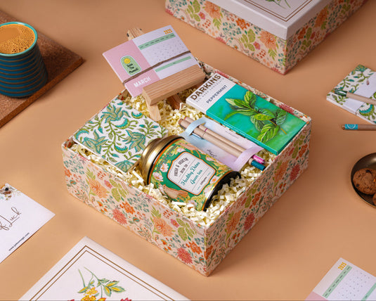 A gift box containing a variety of items including a packet of green tea, a desk calendar, chocolate, a handcrafted tearable desk pad, and eco-friendly seed pencils and pens.