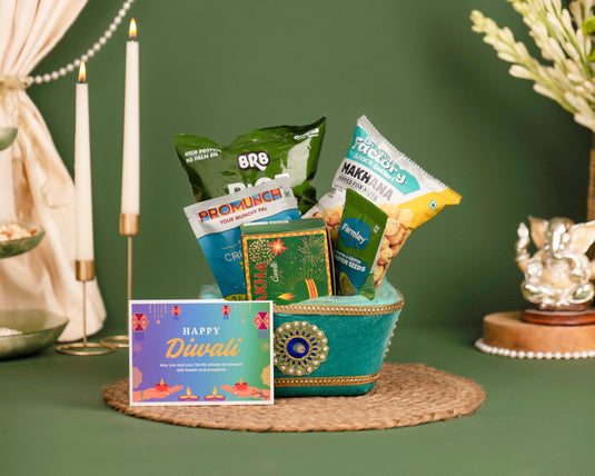 A Diwali gift basket arrangement featuring a teal decorative bowl filled with assorted snacks and a colorful 'Happy Diwali' greeting card, styled on a woven mat against a dark green background with candles and decorative props.